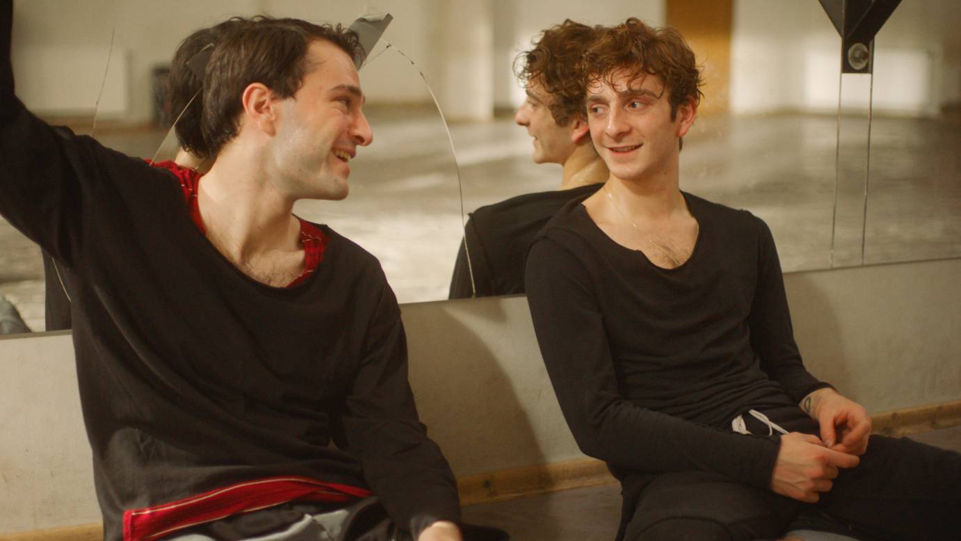 Merak and Irakli smile at each other as they sit against a mirror in a dance studio